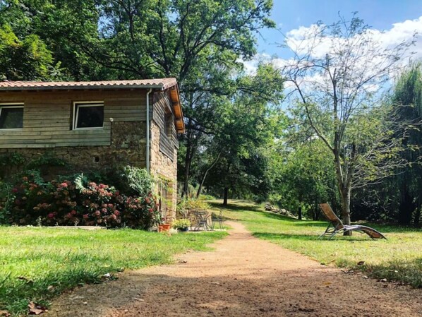 A rustic wooden house with a stone foundation, surrounded by lush greenery and a dirt path leading to a metal chair.