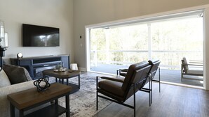 A spacious living room with a large window, a flat-screen TV, and a comfortable seating area. - The Clermont at Clermont Bay (Hancock County)