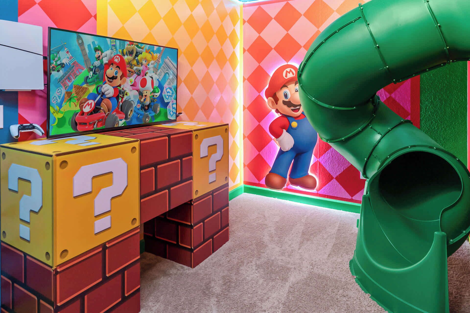 A play area with a Mario-themed mural, a green slide, and a game console on a brick-patterned table.