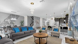 A modern living room with a grey sofa, a round coffee table, and a dining area with a white table and chairs. - Upstay 9BR Villa w Pool & Cinema—Epic Universe (Championsgate)