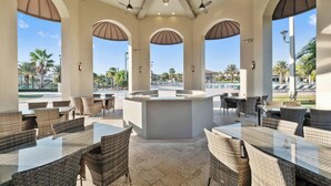An outdoor patio area with wicker furniture, a central bar, and large windows offering views of palm trees and a pool. - Upstay 9BR Villa w Pool & Cinema—Epic Universe (Championsgate)