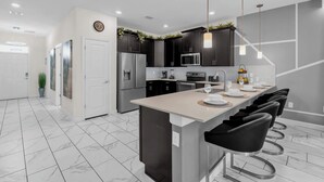 A modern kitchen with dark cabinets, stainless steel appliances, a central island with bar stools, and a white door. - Upstay 9BR Villa w Pool & Cinema—Epic Universe (Championsgate)