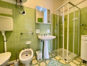 Een badkamer met groene tegels, een witte wastafel, een toilet, een douchecabine van glas en een bidet.