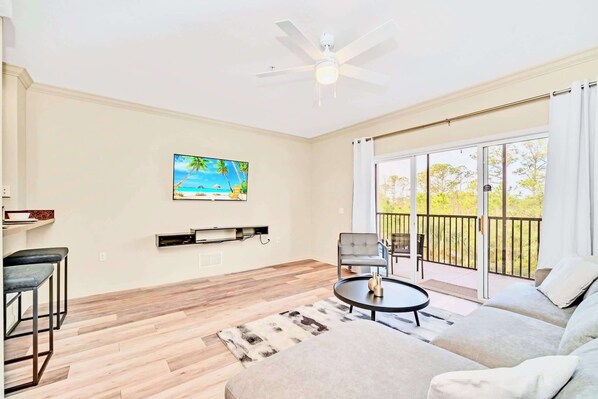 A living room with a sofa, a coffee table, a TV mounted on the wall, and a balcony with a view of trees. - Upstay: Resort Style Condo w Pool, Sauna (Winter Garden)