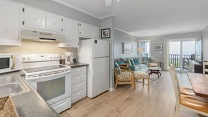 A modern kitchen with white cabinetry, a stove, and a refrigerator. There is a dining area with wooden chairs and a table. The room has a large window with a view of the outdoors. - Seabound - Oceanfront Condo + Steps to the Sand (Atlantic Beach)