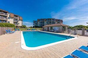A rectangular swimming pool with a white edge, surrounded by a brick patio. There are blue lounge chairs and a small building with a sign. - Seabound - Oceanfront Condo + Steps to the Sand (Atlantic Beach)
