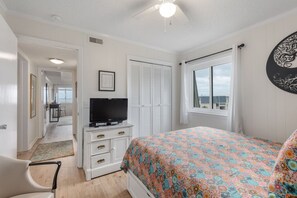 A bedroom with a bed, a TV on a dresser, a window with curtains, and a view of the outside. - Seabound - Oceanfront Condo + Steps to the Sand (Atlantic Beach)