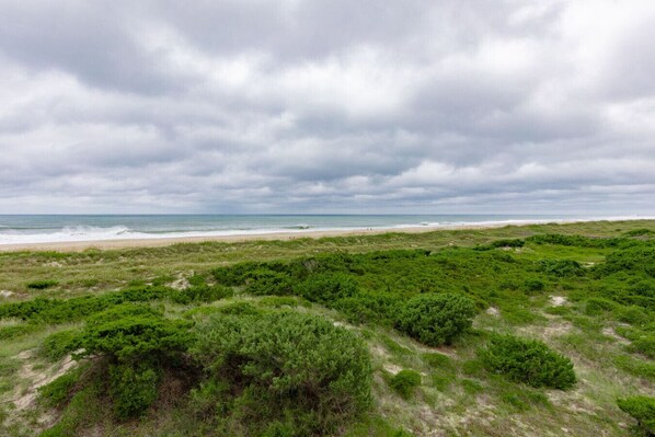 A coastal landscape with a beach, grassy dunes, and a cloudy sky. - Seabound - Oceanfront Condo + Steps to the Sand (Atlantic Beach)