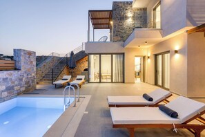A modern outdoor pool area with lounge chairs, a stone wall, and a staircase leading to a balcony.
