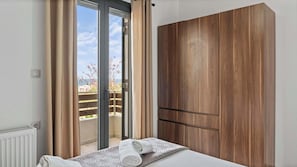 A bedroom with a bed, a wooden wardrobe, a sliding glass door to a balcony, and a view of the outdoors.