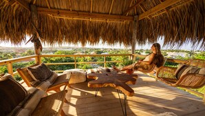 Una terraza de madera con techo de paja, muebles de mimbre y vistas a zonas verdes y edificios.
