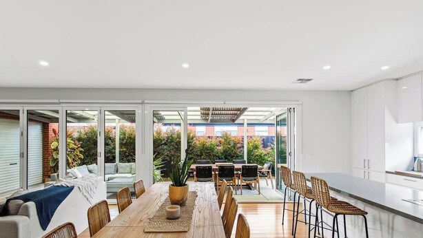 Una cocina moderna con armarios blancos, un comedor con mesa y sillas de madera, y un amplio ventanal corredero que da a un jardín.