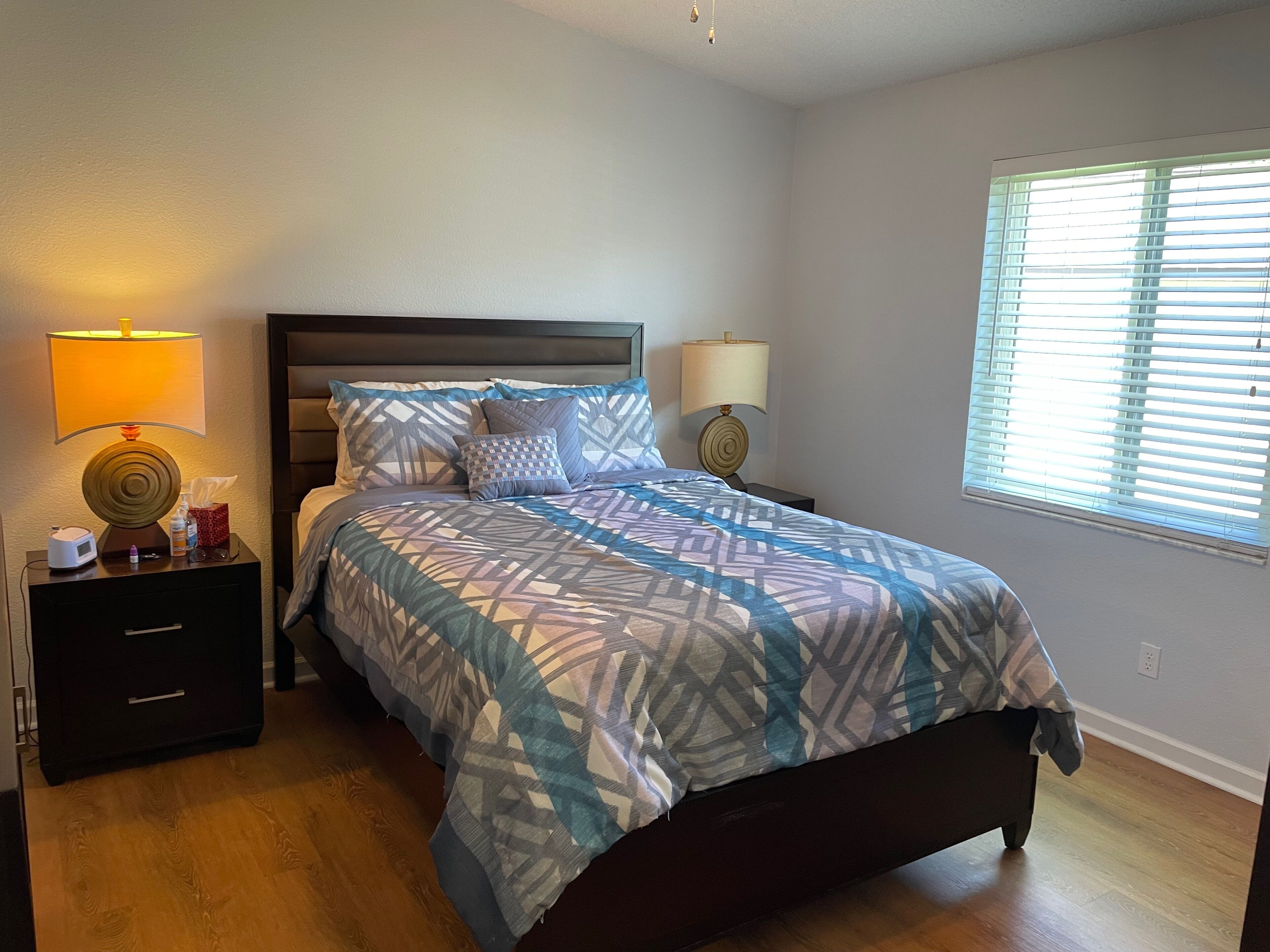 A bedroom with a bed, two bedside tables, lamps, a nightstand, and a window with blinds.