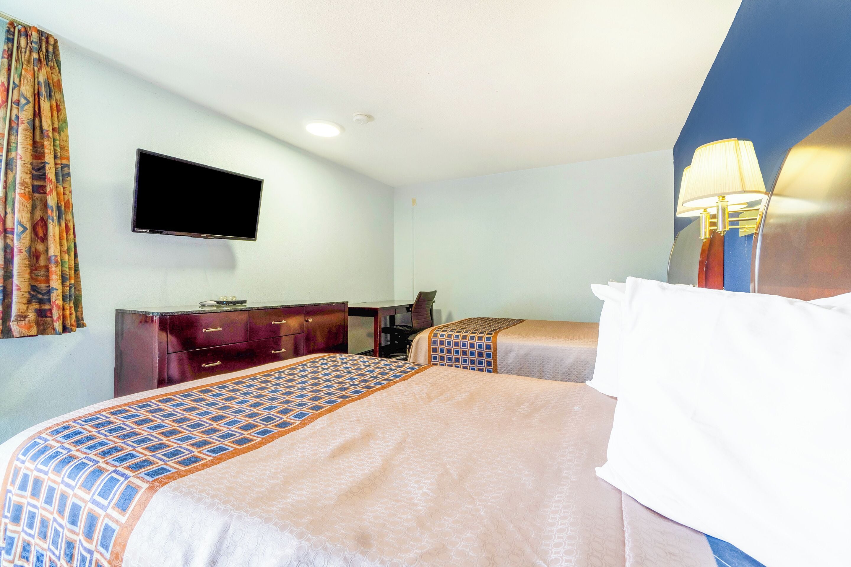 A hotel room with two beds, a TV mounted on the wall, a desk, and a lamp.