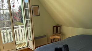 A bedroom with a bed, a chair, a small table, a door leading to a balcony, and a framed picture on the wall.