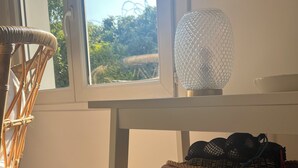 A room with a wicker chair, a wooden crate on a white table, a thermostat, and a window with a view of greenery outside. - Villa 60m² with garden, air conditioning and parking - up to 6 people (La Roquette-sur-Siagne)
