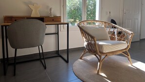 A room with a grey chair, a wicker chair with a cushion, a wooden desk, and a window with a view of greenery. - Villa 60m² with garden, air conditioning and parking - up to 6 people (La Roquette-sur-Siagne)