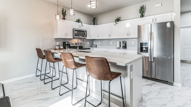 A modern kitchen with white cabinets, stainless steel appliances, and a marble countertop. There are four bar stools and pendant lights above the island. - Serene Golf View Retreat 3 Bedroom Duplex by RedAwning (Lehigh Acres)
