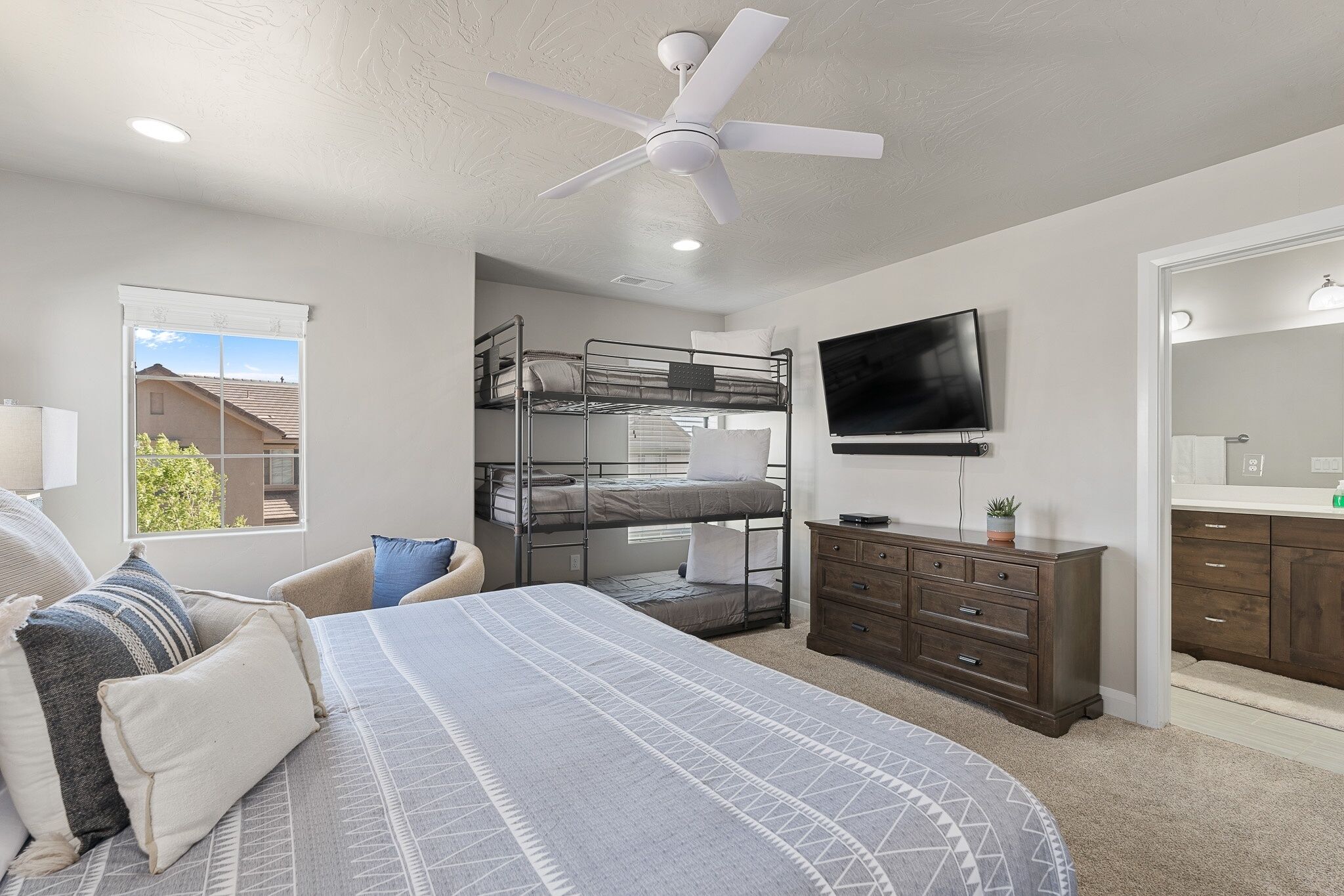 A bedroom with bunk beds, a flat-screen TV, a ceiling fan, a dresser, and a window with a view of houses.