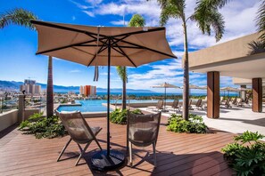 A rooftop pool area with lounge chairs, a large umbrella, and a view of the ocean and cityscape. - Top-Floor 2BR w/ Skyline Views, Pool & Gym (Puerto Vallarta)