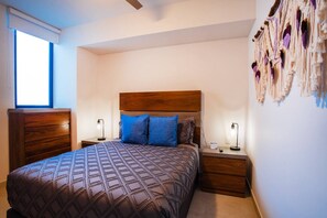 A bedroom with a wooden headboard, a patterned bedspread, two nightstands with lamps, and a wall-mounted macrame decoration. - Top-Floor 2BR w/ Skyline Views, Pool & Gym (Puerto Vallarta)