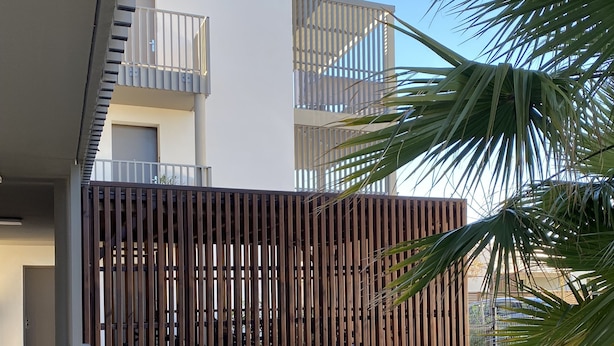 A modern building with a balcony, a wooden fence, and a small garden area with plants and a blue mat. - Appart 4 Pers Climatisé Avec Terrasse, Parking et Piscine Collective - V0816 (Vias)