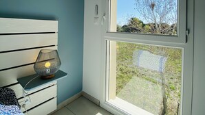 A room with a large window, a bed, a bedside table with a lamp, and a wall-mounted decorative item. - Appart 4 Pers Climatisé Avec Terrasse, Parking et Piscine Collective - V0816 (Vias)