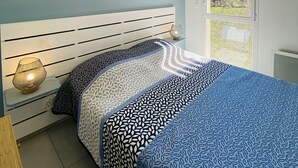 A bedroom with a bed, a nightstand, a wall-mounted light fixture, and a window with a view of trees. - Appart 4 Pers Climatisé Avec Terrasse, Parking et Piscine Collective - V0816 (Vias)