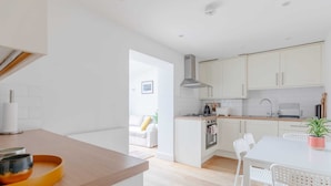 A modern kitchen with white cabinets, a central island, and a dining area with white chairs. - Homely 2BD Flat W/private Patio Near Maida Vale! (London)