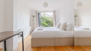 A modern bedroom with a large bed, a small table, and a view of greenery outside. - Homely 2BD Flat W/private Patio Near Maida Vale! (London)