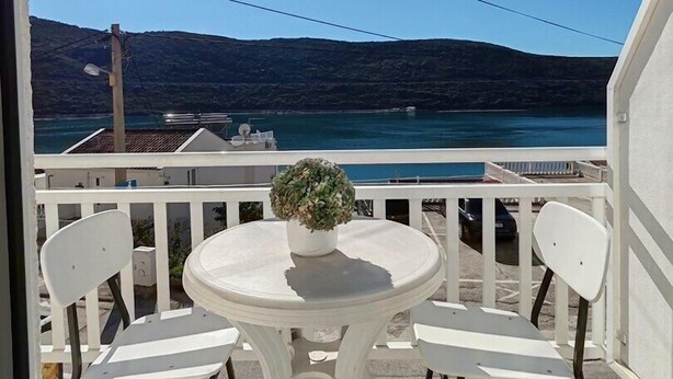 A balcony with a round table and two chairs, overlooking a coastal area with a clear sky and distant hills.