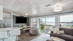Un salon moderne avec un coin cuisine, un grand téléviseur et un balcon offrant une vue sur l’eau.