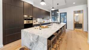 Een moderne keuken met donkerhouten kastjes, roestvrijstalen apparatuur, een marmeren eiland met barstoelen en hanglampen.