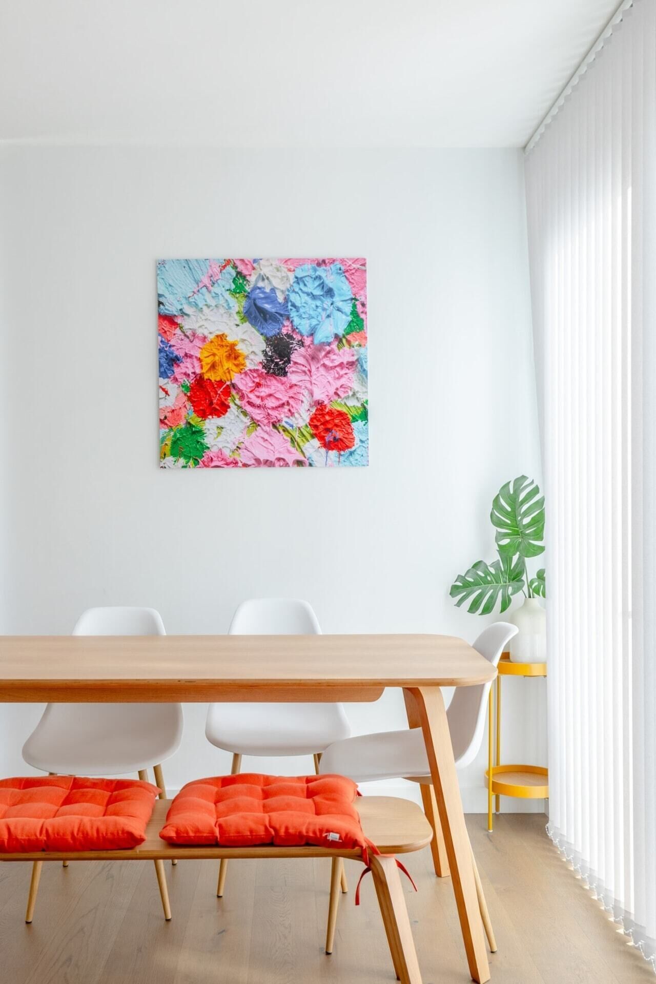 A dining area with a wooden table, white chairs, and orange cushions. A colorful abstract painting is on the wall, and a large green plant is in the corner.