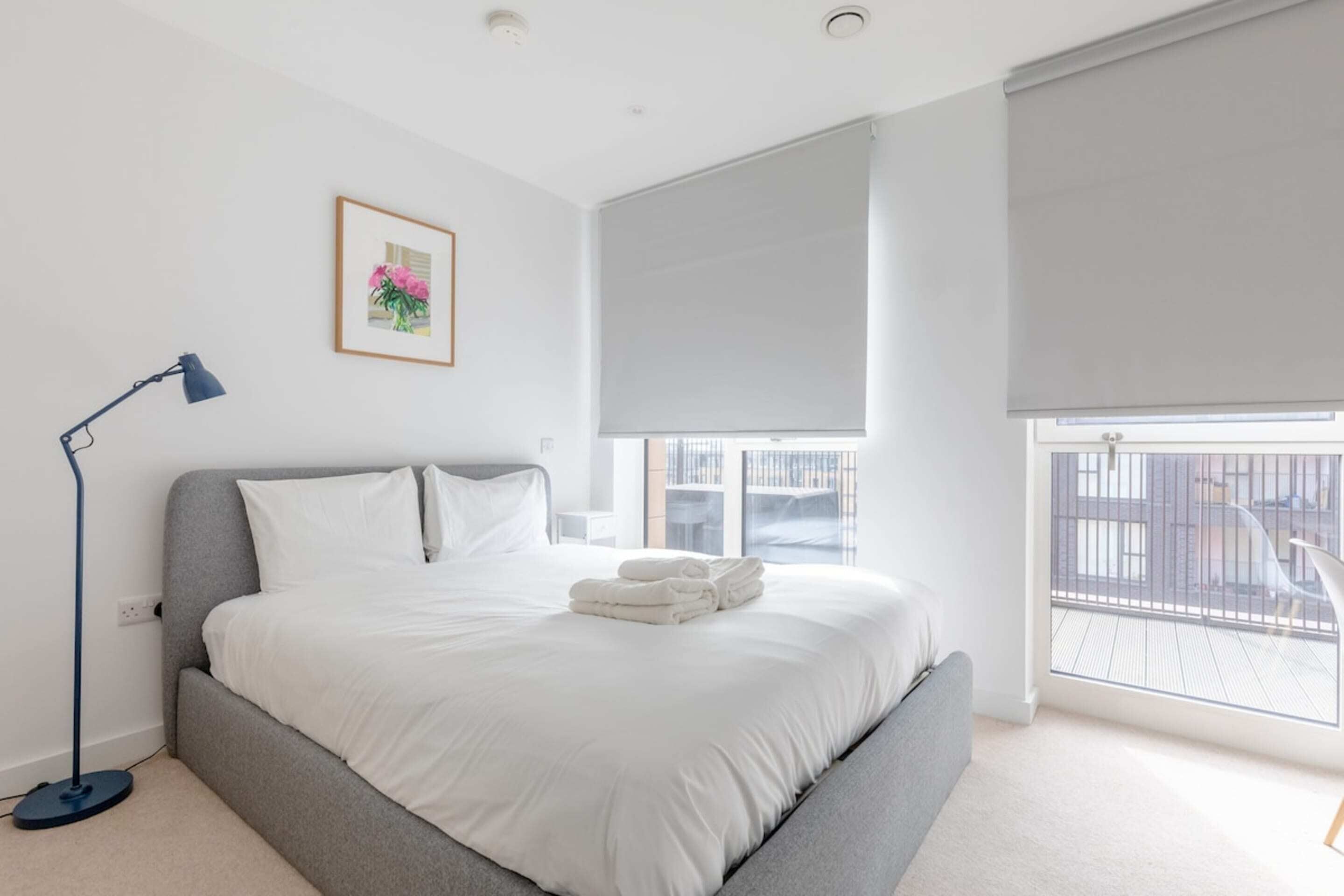 Une chambre moderne avec un grand lit, une lampe sur pied, une fenêtre avec des stores et un tableau encadré au mur.