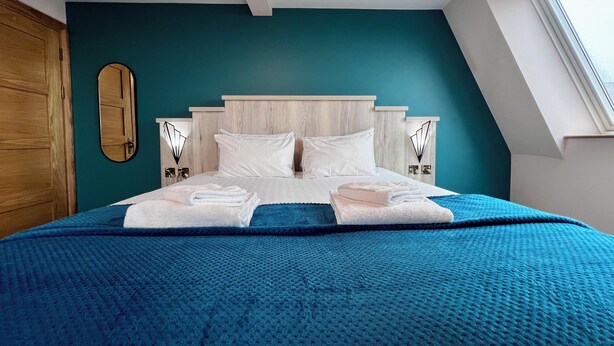 A neatly made bed with white linens and a blue blanket, a wooden headboard, and a teal accent wall. - The Bath Rooms (Morecambe)