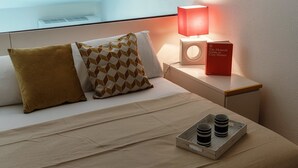 Una cama bien tendida con dos almohadas, una mesita de noche con una lámpara y un libro, y una bandeja con dos tazas.