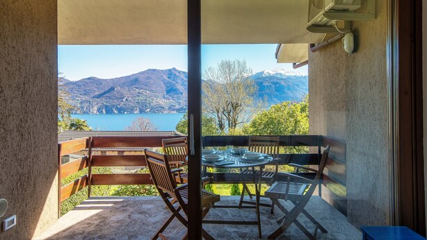 Un balcón con mesa y sillas, con vista a un paisaje montañoso y una masa de agua.