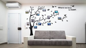 A room with a grey sofa, a wall decorated with a tree and photos, and a white door. - House in the historic center near the port (Trapani)