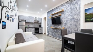A modern living room with a sofa, dining table, and chairs. A kitchen with a stove, oven, and cabinets is visible in the background. - House in the historic center near the port (Trapani)