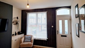 A room with a white door, a window with vertical blinds, a light fixture, a small sofa, and a wall-mounted shelf with decorative items. - Fantastic Home! Football Fans -5 min to Stadium (Liverpool)
