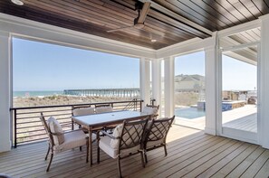 Uma área externa coberta com deck de madeira, uma mesa com cadeiras e vista para a praia e o oceano.