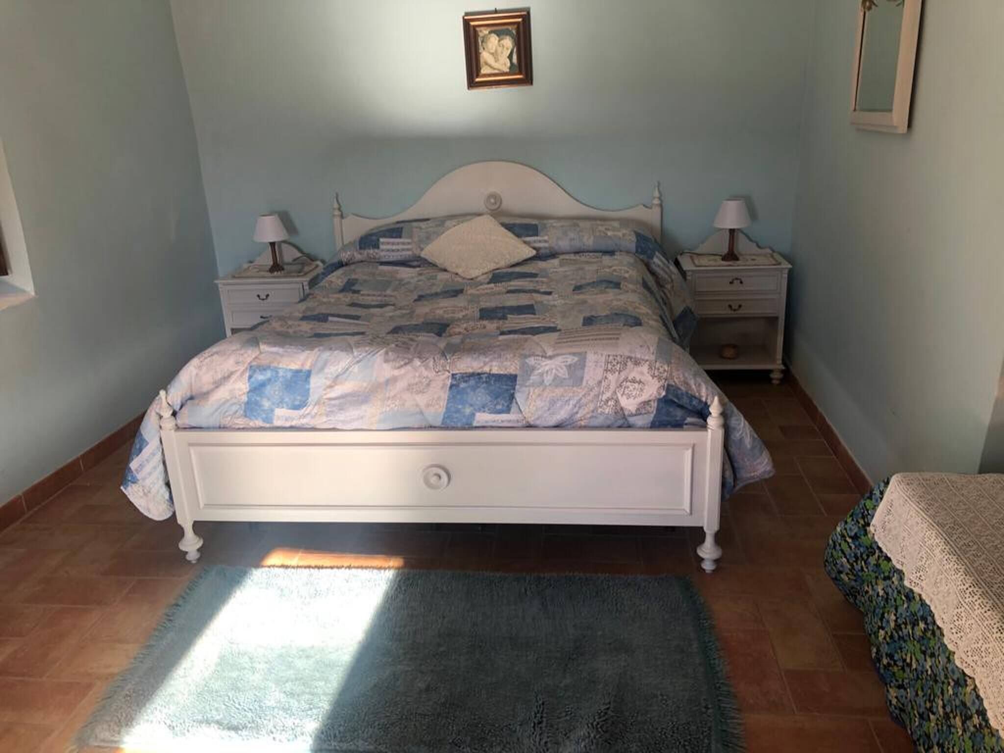 Un dormitorio con una cama, dos mesitas de noche, una mesita auxiliar, una alfombra y un cuadro enmarcado en la pared.