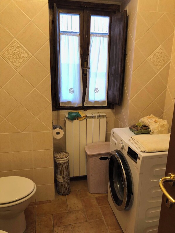 A compact bathroom with a toilet, a washing machine, a radiator, and a window with curtains.