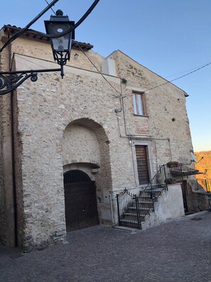 A historic stone building with a central arched doorway, a small window, and a staircase with a metal railing.