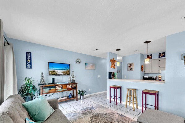 Una sala de estar con un sofá, una mesa de centro y un televisor. Hay una zona de cocina con taburetes y una encimera.