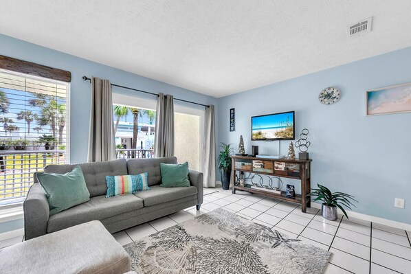 A living room with a grey sofa, a coffee table, a TV on a wooden stand, and a view of the outdoors through large windows. - 🌟 50Ft from Beach Access- Pool View- Budget Friendly- 1BR Nautical Watch E-3🌟 (Panama City Beach)