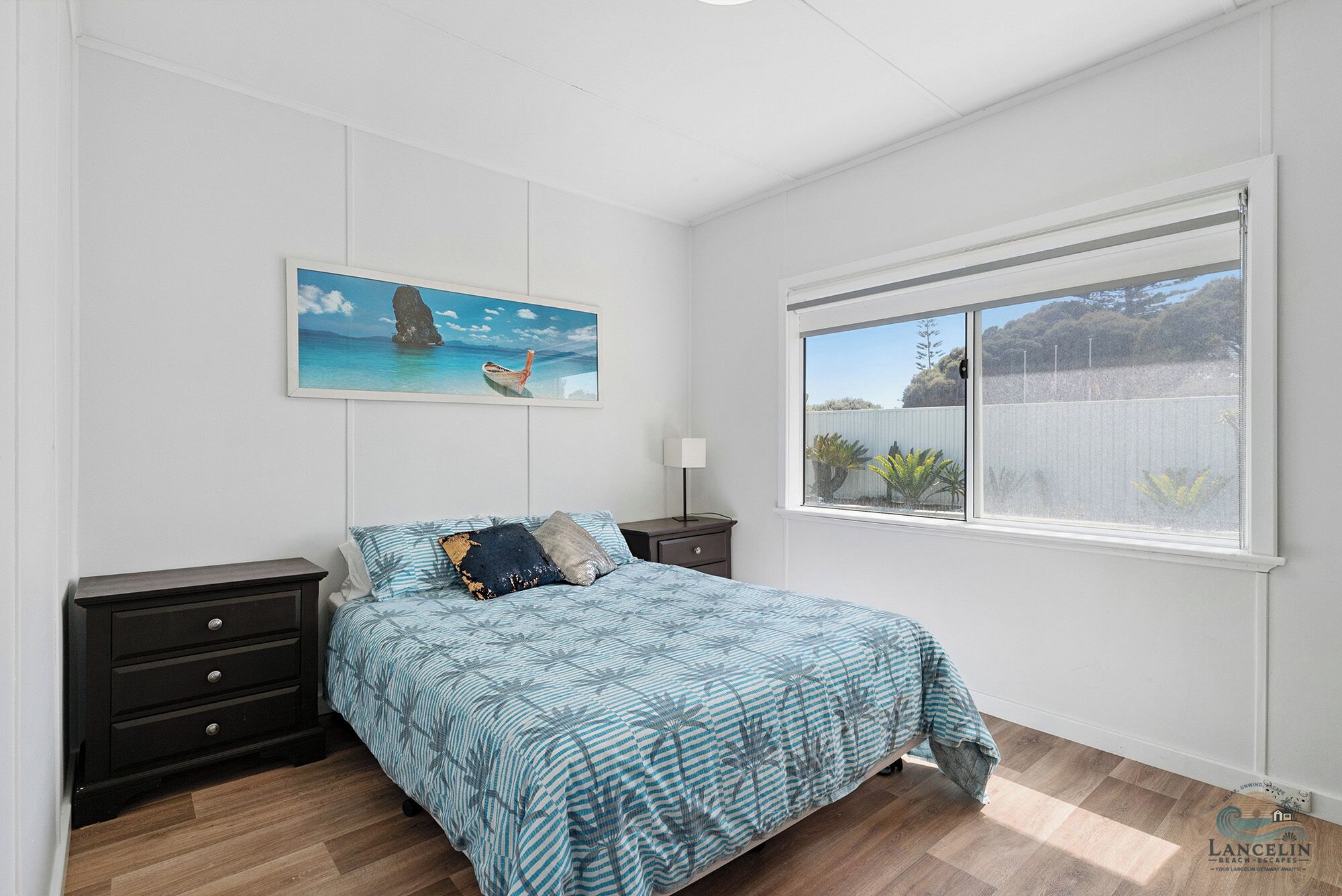 A bedroom with a bed, a nightstand, a lamp, a window with a view, and a painting on the wall.