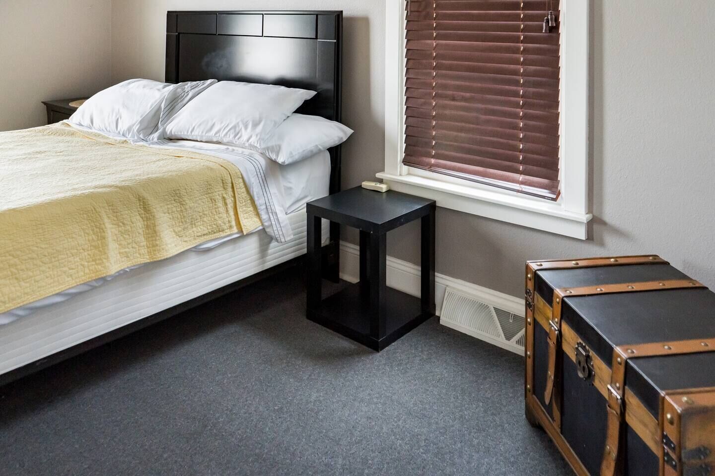 Un dormitorio con cama, mesita de noche, ventana con persianas y un baúl.