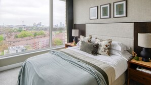 A hotel room with a large window offering a city view, a neatly made bed with multiple pillows, and a bedside table with a lamp and a small vase. - Designer Penthouse Panoramic Views London 2 Beds (London)
